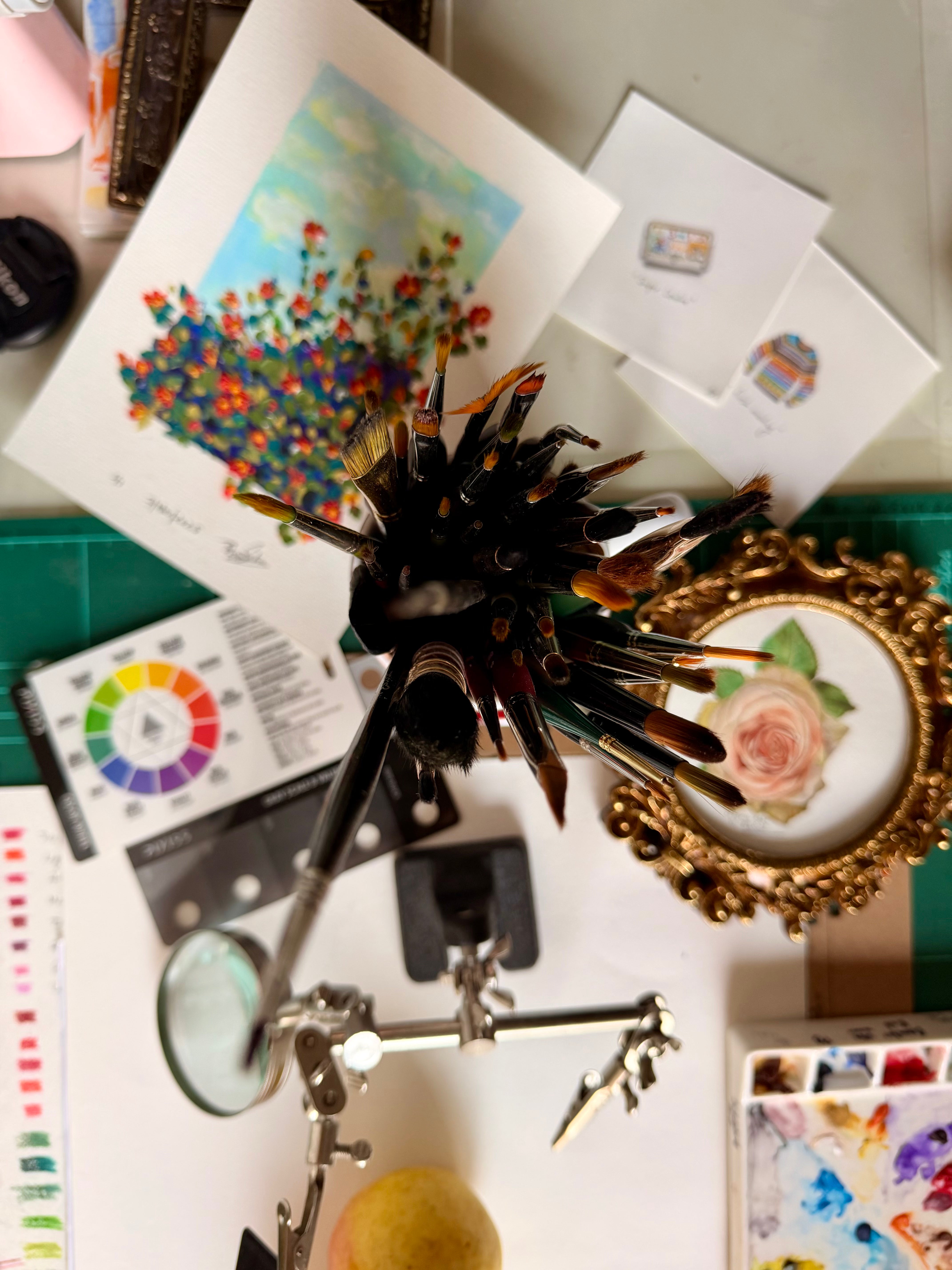 A top-down view of Artist Bhawana Sahu's creative workspace featuring a central paintbrush holder filled with multicolored brushes, surrounded by art supplies including a watercolor painting of a colorful tree, a small framed rose illustration, a color wheel, and a paint palette.